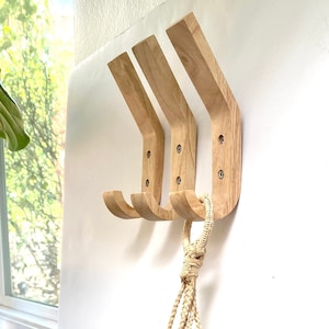 May include: Three wooden wall hooks with a curved design are mounted on a white wall. A hanging plant in a woven basket is suspended below the hooks. The plant has green and white leaves.