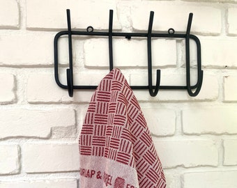 May include: A black metal hook rack with five hooks mounted on a white brick wall. A red and white striped kitchen towel with the text "UR IAP & REL EQU" is hanging on one of the hooks.