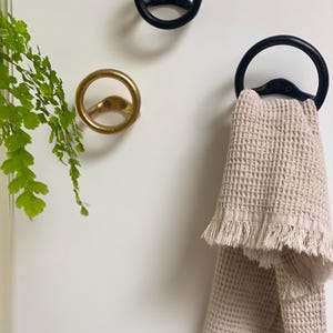 Puede incluir: Dos ganchos de toalla de metal negro con un dise&ntilde;o circular. Un gancho tiene una toalla colgada. La toalla es de color beige claro con un borde de flecos.