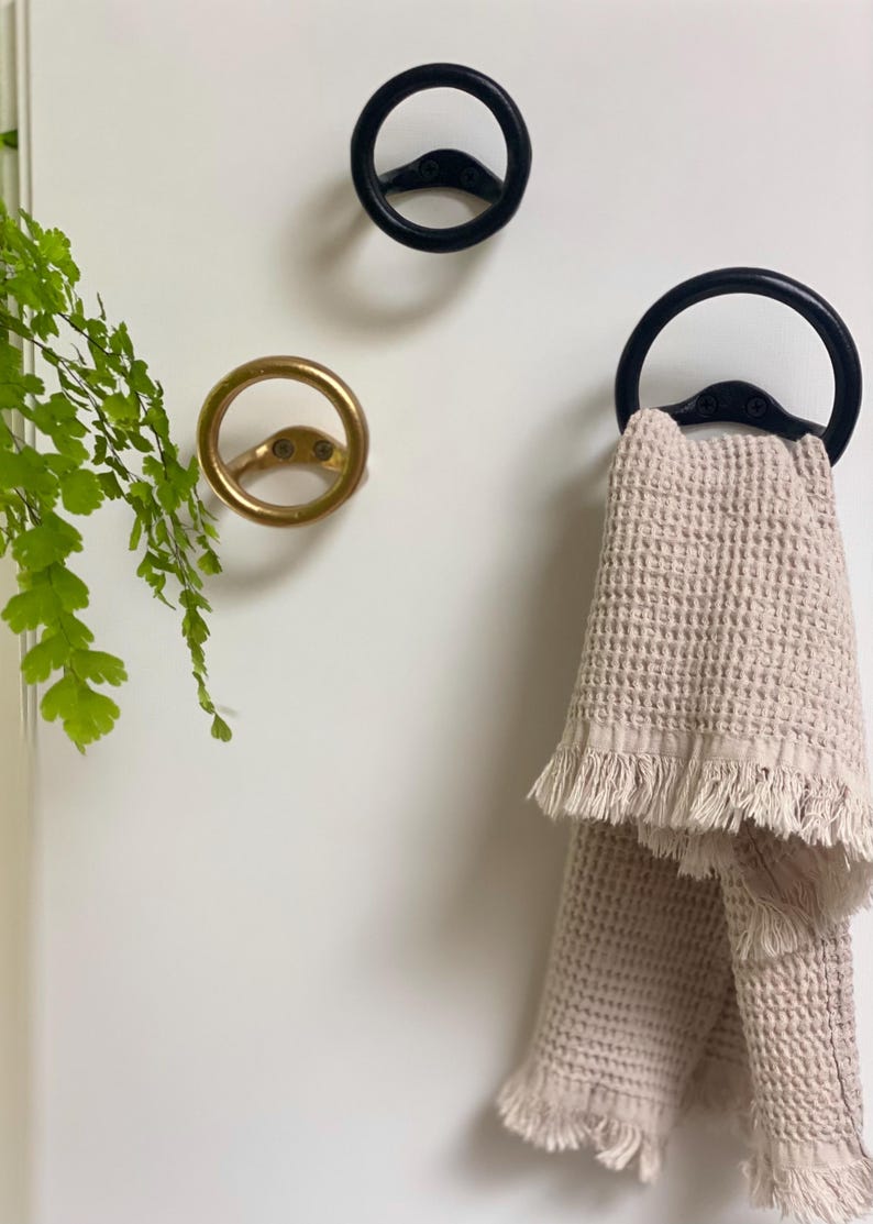 Puede incluir: Dos ganchos de toalla de metal negro con un dise&ntilde;o circular. Un gancho tiene una toalla colgada. La toalla es de color beige claro con un borde de flecos.