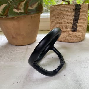 Puede incluir: Un soporte para anillos de metal negro con forma redondeada. El soporte est&aacute; dise&ntilde;ado para sujetar un anillo de forma segura.