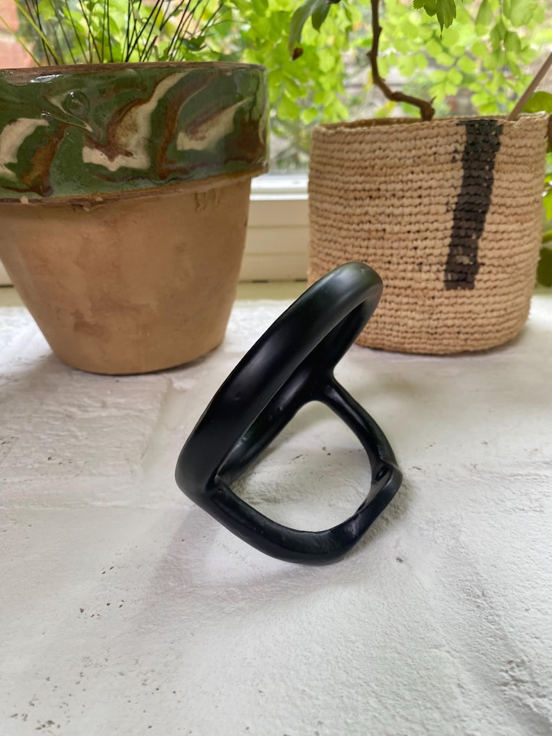 Puede incluir: Un soporte para anillos de metal negro con forma redondeada. El soporte est&aacute; dise&ntilde;ado para sujetar un anillo de forma segura.