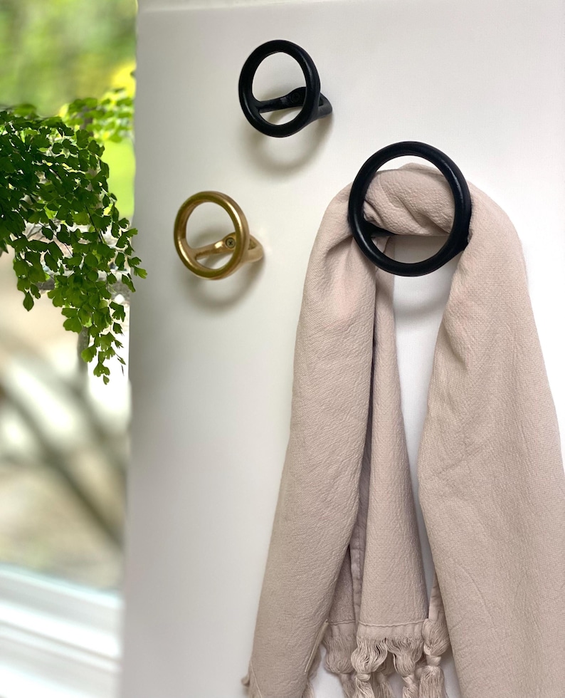 Puede incluir: Tres ganchos de pared decorativos con forma de c&iacute;rculo. Dos ganchos son negros y uno es dorado. Una bufanda marr&oacute;n claro est&aacute; colgada en uno de los ganchos negros.