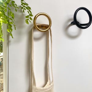 Puede incluir: Dos ganchos de pared circulares, uno dorado y otro negro, est&aacute;n montados en una pared blanca. Una bolsa de cuerda blanca cuelga del gancho dorado.