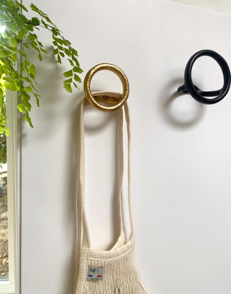 Puede incluir: Dos ganchos de pared circulares, uno dorado y otro negro, est&aacute;n montados en una pared blanca. Una bolsa de cuerda blanca cuelga del gancho dorado.