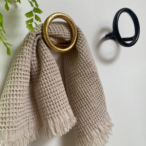 Puede incluir: Una toalla de tejido de waffle beige con flecos colgada de un gancho de anillo dorado. Tambi&eacute;n se ve un gancho de anillo negro en la pared.