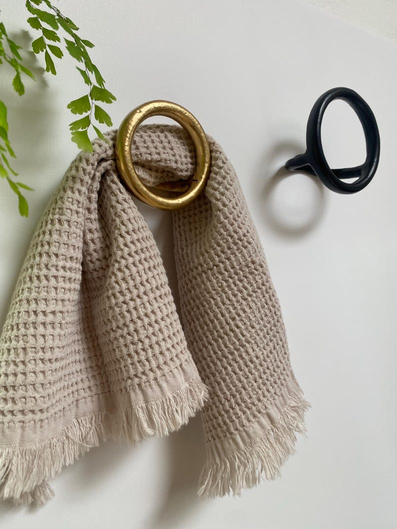 Puede incluir: Una toalla de tejido de waffle beige con flecos colgada de un gancho de anillo dorado. Tambi&eacute;n se ve un gancho de anillo negro en la pared.