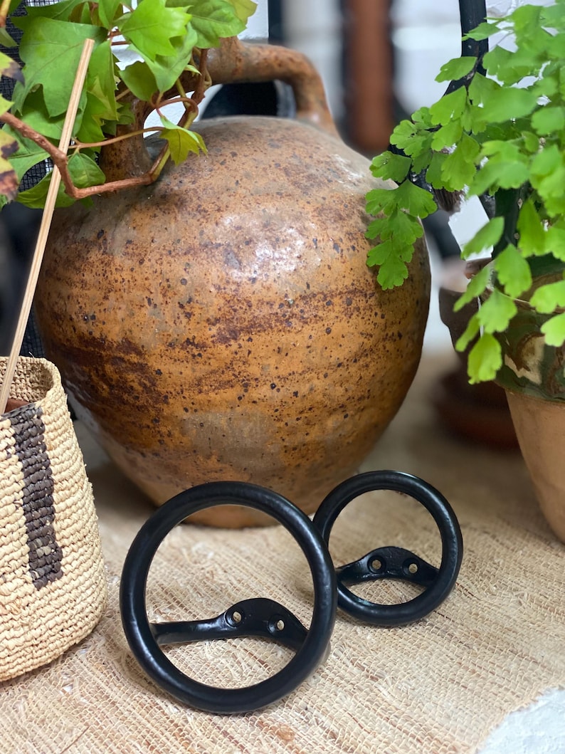 Puede incluir: Dos ganchos de metal negro con un dise&ntilde;o circular. Los ganchos est&aacute;n colgados en una superficie tejida.