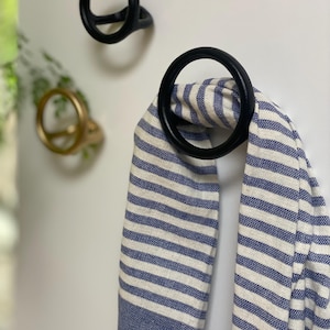 Puede incluir: Dos ganchos de metal negro con un dise&ntilde;o circular. Un gancho tiene una toalla a rayas azules y blancas colgada.