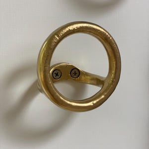 Puede incluir: Gancho de metal dorado con un dise&ntilde;o circular. El gancho est&aacute; fijado a una pared con dos tornillos.