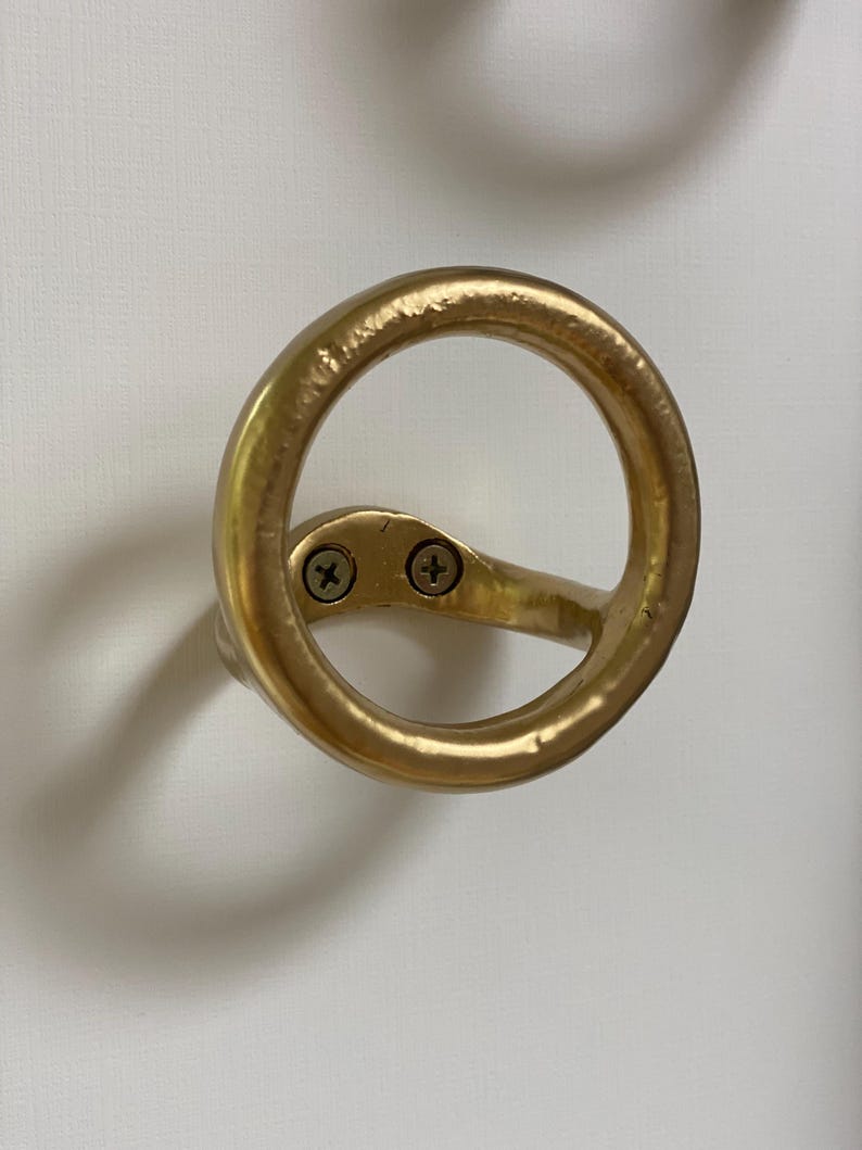 Puede incluir: Gancho de metal dorado con un dise&ntilde;o circular. El gancho est&aacute; fijado a una pared con dos tornillos.