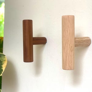 May include: Two wooden T-shaped hooks, one brown and one natural wood, mounted on a white wall.
