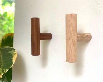 May include: Two wooden T-shaped hooks, one brown and one natural wood, mounted on a white wall.
