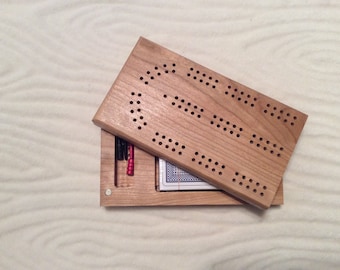 May include: Wooden cribbage board with a hinged lid, featuring a light brown finish. The board has multiple rows of drilled holes for pegging and a compartment for storing pegs and a deck of cards. The pegs are red and black.
