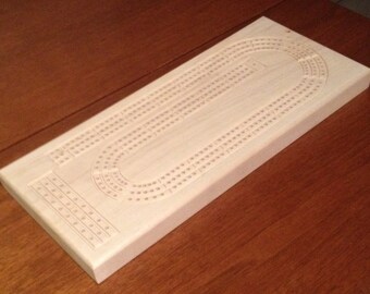May include: A wooden cribbage board with a natural finish. The board has a traditional design with a large oval track and numbered squares.