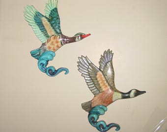 May include: Two decorative wall art pieces depicting ducks in flight. The ducks have detailed wings with shades of green, brown, and orange. Each duck has a red beak and is attached to a blue wave design.