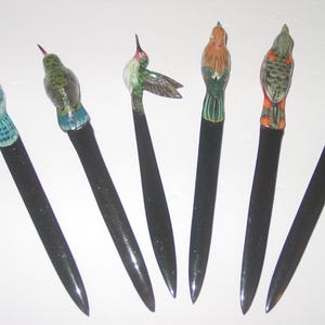 May include: Six letter openers with bird figurines. Each has a long, black handle and a different bird design, painted in colours like blue, green, orange, and red. The birds have detailed feather patterns.