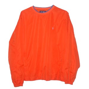 May include: A bright orange long-sleeve pullover windbreaker jacket featuring a crew neckline with a thin blue trim. The garment has a small white embroidered logo on the left chest and elasticized cuffs at the end of each long sleeve.