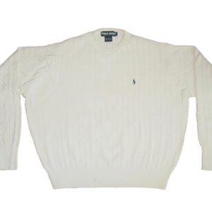 May include: A cream-colored long-sleeve cable knit sweater featuring a crew neckline and ribbed cuffs and hem. The garment displays a small embroidered Polo Golf logo on the left chest and a Polo Golf brand label at the interior back neckline.