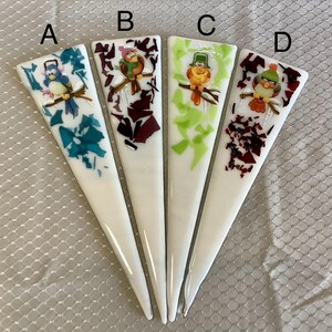 May include: Four white, triangular-shaped decorative pieces with cartoon bird designs. Each piece features a different bird with colorful accents and a unique background of teal, burgundy, or green. The birds are perched on branches.