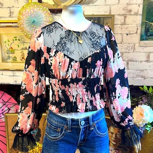 May include: A black floral print blouse with pink flowers and a lace neckline is paired with denim shorts. The blouse has long sleeves with black lace cuffs. A gold necklace with a heart pendant is visible.