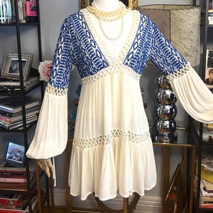 Pu&ograve; includere: Un abito color crema con dettagli ricamati blu sul corpetto e sulle maniche. L'abito presenta uno scollo a V, maniche lunghe e fluide e una gonna a balze. Un cappello di paglia con una fascia nera e una collana di perle completano il look. L'abito &egrave; esposto su un manichino.