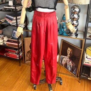 May include: A mannequin displays a black mesh crop top and high-waisted red pants. The outfit is accessorized with a black hat with a gold band and bracelets. The pants are pleated and have a wide leg. The mannequin is on a rolling stand.