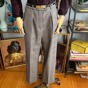 Puede incluir: Un maniquí exhibe una blusa burdeos con mangas abullonadas y pantalones de cuadros plisados. Un cinturón negro con hebilla dorada ciñe la cintura. Los pantalones son grises con un sutil estampado de cuadros rojos y negros. El conjunto está sobre un soporte rodante.