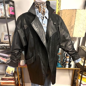 May include: A black leather jacket with textured panels on the shoulders and upper sleeves. The jacket has a wide collar, a button closure, and two front pockets. The jacket is paired with a denim shirt and blue jeans.