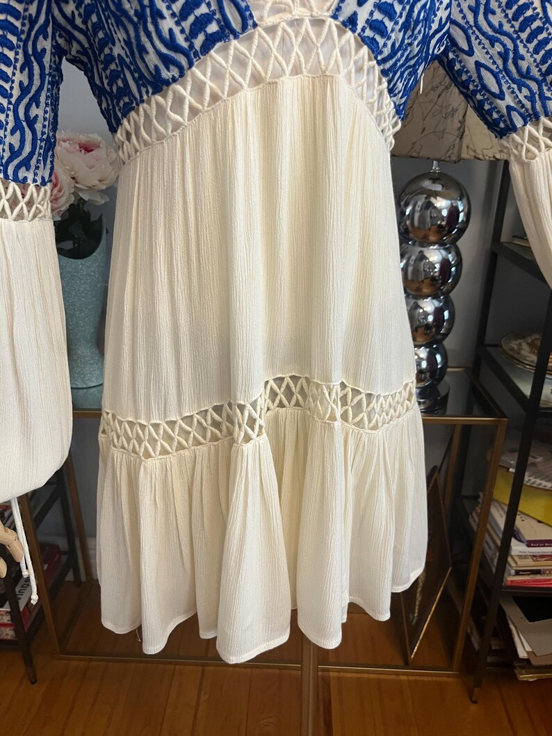Pu&ograve; includere: Un vestito color crema con maniche e corpetto ricamati in blu. L'abito presenta un design all'uncinetto sullo scollo e in vita, e una gonna fluida. Le maniche sono lunghe e ampie, e l'abito &egrave; realizzato in un tessuto leggero.