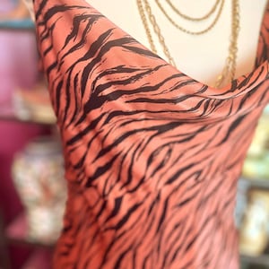 Puede incluir: Un vestido color melocot&oacute;n con un estampado de rayas de tigre negras. El vestido tiene un cuello de cisne y se exhibe en un maniqu&iacute;. Tambi&eacute;n se ven collares dorados con un relicario.
