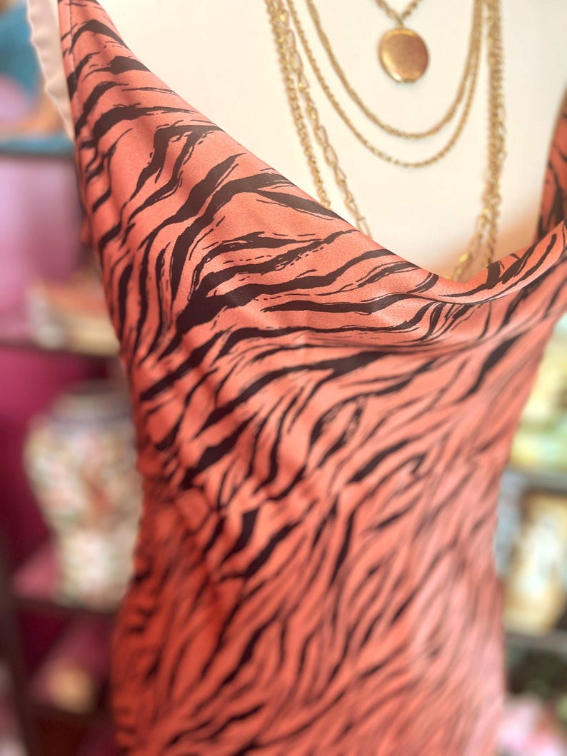 Puede incluir: Un vestido color melocot&oacute;n con un estampado de rayas de tigre negras. El vestido tiene un cuello de cisne y se exhibe en un maniqu&iacute;. Tambi&eacute;n se ven collares dorados con un relicario.