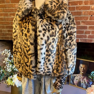 K&ouml;nnte beinhalten: Eine kurze Kunstfelljacke mit braun-schwarzem Leopardenmuster, einem breiten Kragen und einem einzelnen schwarzen Knebelverschluss am Halsausschnitt. Die Jacke wird &uuml;ber einem passenden lang&auml;rmeligen Oberteil und einer hellblauen Acid-Wash-Jeans getragen.