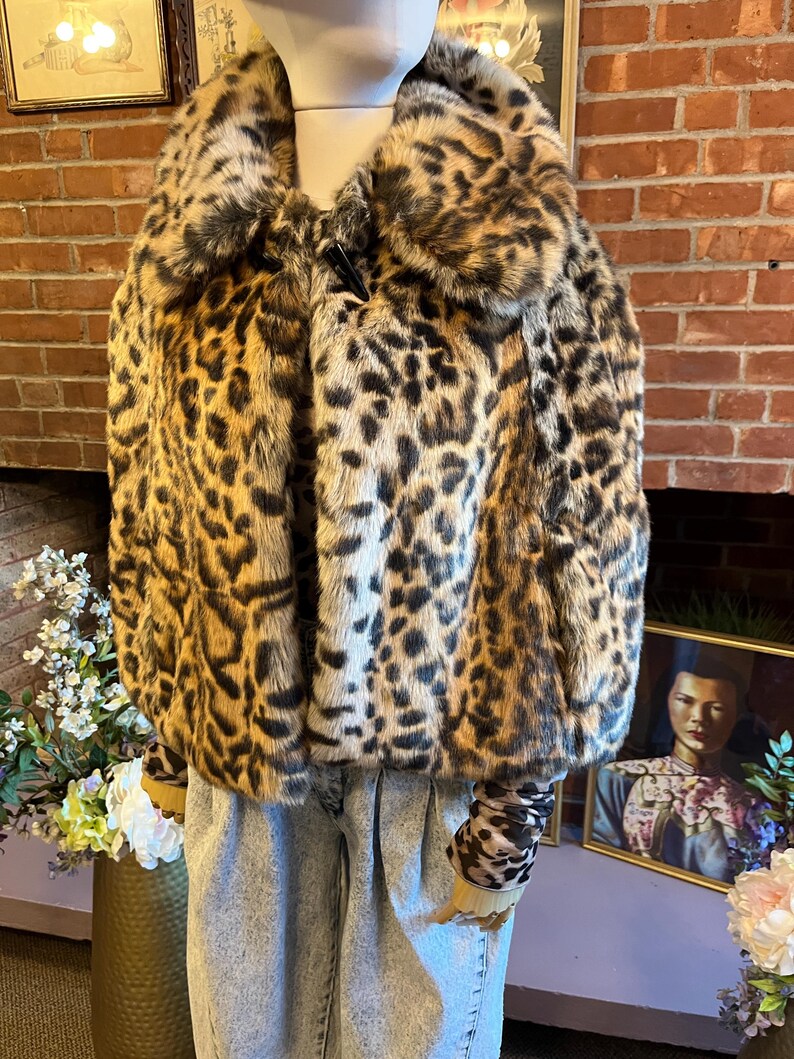 K&ouml;nnte beinhalten: Eine kurze Kunstfelljacke mit braun-schwarzem Leopardenmuster, einem breiten Kragen und einem einzelnen schwarzen Knebelverschluss am Halsausschnitt. Die Jacke wird &uuml;ber einem passenden lang&auml;rmeligen Oberteil und einer hellblauen Acid-Wash-Jeans getragen.