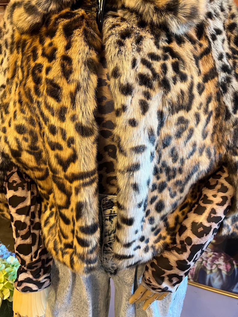 K&ouml;nnte beinhalten: Eine kurze Kunstfelljacke mit einem braunen, schwarzen und beigen Leopardenmuster. Die Jacke wird &uuml;ber einem lang&auml;rmeligen Oberteil mit passendem Leopardenmuster und einer hellblauen, s&auml;uregewaschenen Jeans mit sichtbarem Knopf getragen.