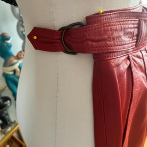 May include: A close-up of a red leather belt with a dark metal buckle. The belt is wrapped around a mannequin. The leather has a textured appearance and is stitched along the edges. A yellow pin is placed on the belt.