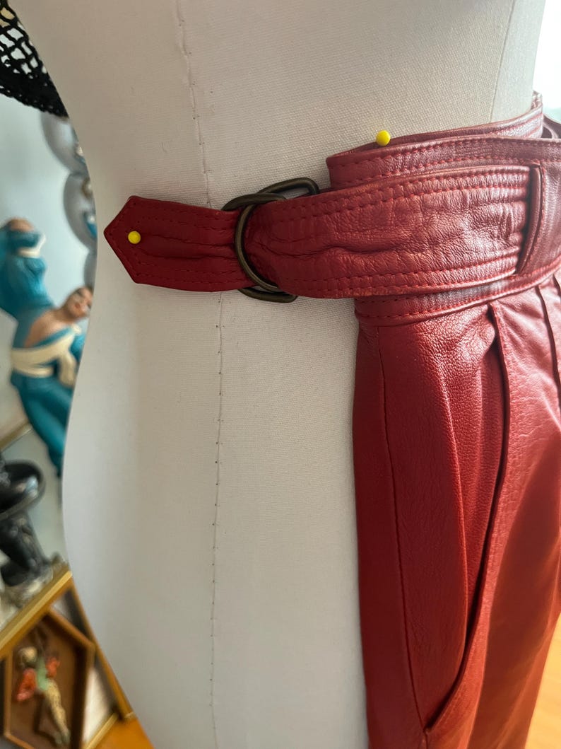 May include: A close-up of a red leather belt with a dark metal buckle. The belt is wrapped around a mannequin. The leather has a textured appearance and is stitched along the edges. A yellow pin is placed on the belt.