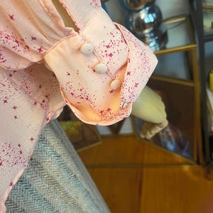 Może przedstawiać: Zbliżenie na jasnor&oacute;żowy rękaw bluzki z wzorem gwiazd i plamek w ciemniejszym r&oacute;żu. Mankaet ma trzy małe, okrągłe, białe guziki. Tkanina wydaje się być lekkim materiałem.