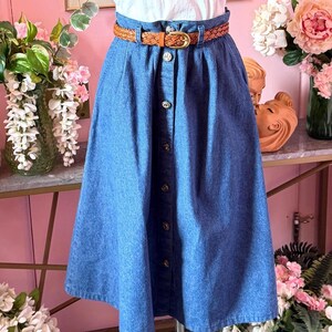 May include: A high-waisted blue denim midi skirt featuring a vertical row of silver-toned metal buttons down the center front. The skirt has a gathered waistline and is cinched with a woven brown leather belt that has a gold-toned metal buckle.