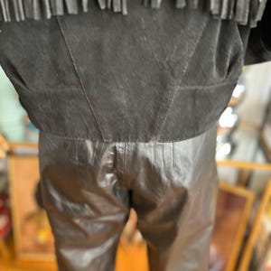 May include: Black leather pants and a black suede jacket with fringe detail. The jacket has a cropped fit, and the pants appear to be high-waisted. The outfit is likely vintage or inspired by Western wear.