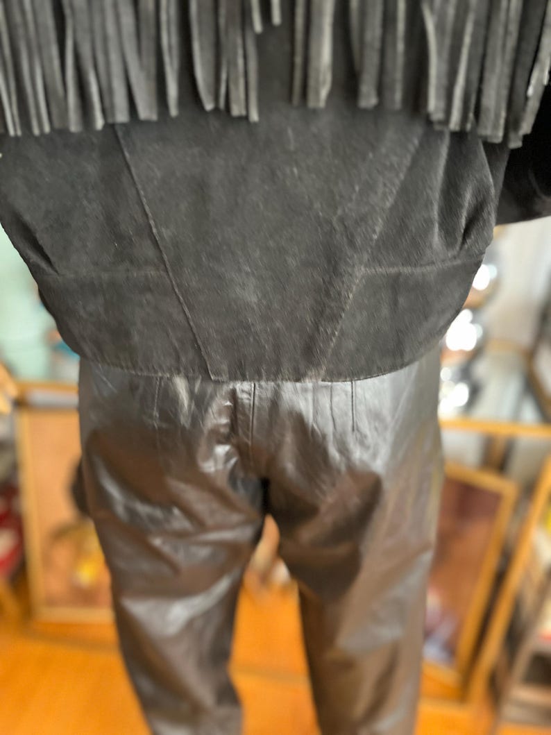 May include: Black leather pants and a black suede jacket with fringe detail. The jacket has a cropped fit, and the pants appear to be high-waisted. The outfit is likely vintage or inspired by Western wear.
