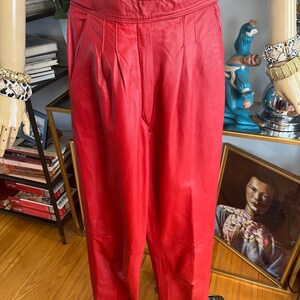 May include: Bright red leather pants with a high waist and pleated front. The pants are displayed on a mannequin. The pants have a classic, tailored design.