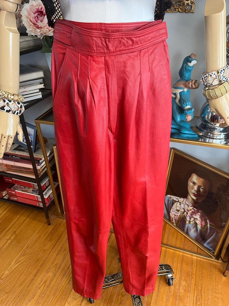 May include: Bright red leather pants with a high waist and pleated front. The pants are displayed on a mannequin. The pants have a classic, tailored design.