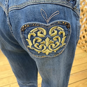 May include: Blue denim jeans with a decorative apple design on the back pocket. The apple is embroidered with gold thread and features a detailed, ornate pattern. The jeans have a classic five-pocket design and are made of a durable denim fabric.