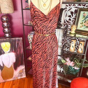 Puede incluir: Un vestido largo color coral con un estampado de rayas de cebra negras. El vestido tiene tirantes finos, un cuello drapeado y un cintur&oacute;n dorado. Un maniqu&iacute; exhibe el vestido, adornado con collares dorados superpuestos.