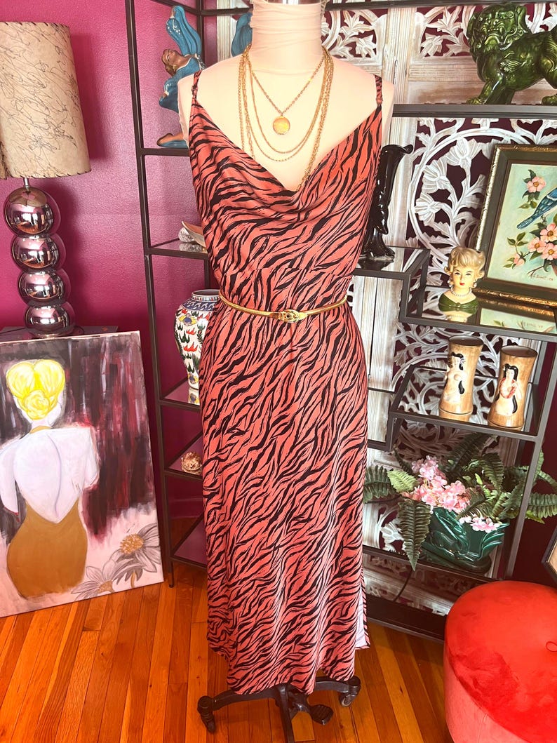 Puede incluir: Un vestido largo color coral con un estampado de rayas de cebra negras. El vestido tiene tirantes finos, un cuello drapeado y un cintur&oacute;n dorado. Un maniqu&iacute; exhibe el vestido, adornado con collares dorados superpuestos.