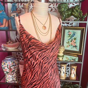 Puede incluir: Un vestido color coral con un estampado de rayas de cebra negras se exhibe en un maniqu&iacute;. El vestido tiene tirantes finos y un cuello drapeado. Un collar dorado en capas est&aacute; drapeado sobre el maniqu&iacute;. El vestido es parte de una colecci&oacute;n de ropa vintage.