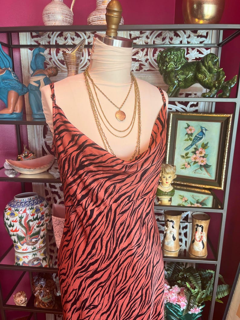 Puede incluir: Un vestido color coral con un estampado de rayas de cebra negras se exhibe en un maniqu&iacute;. El vestido tiene tirantes finos y un cuello drapeado. Un collar dorado en capas est&aacute; drapeado sobre el maniqu&iacute;. El vestido es parte de una colecci&oacute;n de ropa vintage.