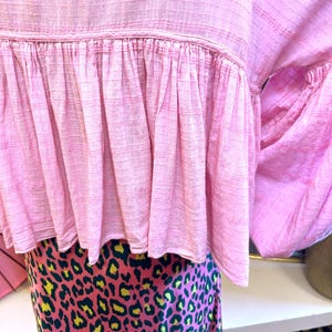 May include: A light pink top with a ruffled hem and a patterned skirt. The top has a textured, grid-like pattern. The skirt features a pink background with a leopard print in yellow and dark green.