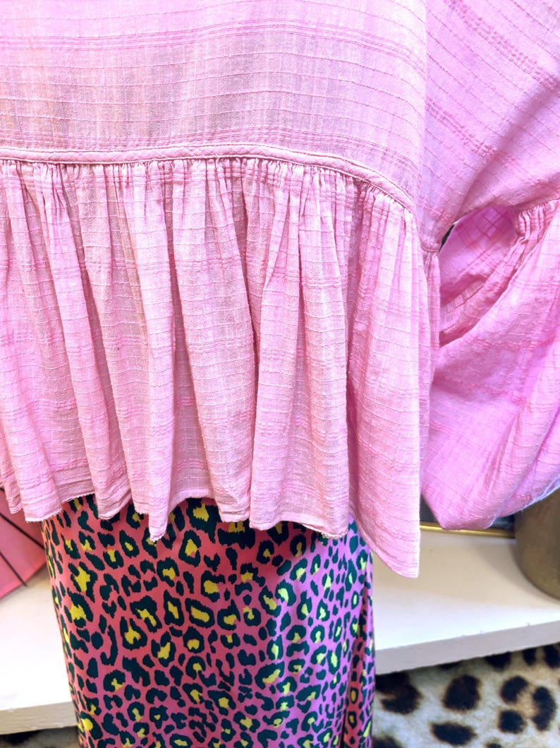 May include: A light pink top with a ruffled hem and a patterned skirt. The top has a textured, grid-like pattern. The skirt features a pink background with a leopard print in yellow and dark green.